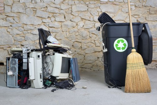 A pile of discarded old computer components, including keyboards, monitors, and various electronic parts, are scattered across a concrete surface in front of a beige stone wall. The items appear mixed, with some cables and small debris around them, indicating they have been recently discarded or awaiting collection. To the right, a large black rubbish bin with a green recycling symbol on its side is positioned upright, holding a few items like a black plastic tray. Leaning against the bin is a traditional straw broom with a wooden handle. The scene likely depicts a rubbish removal situation, possibly outside a property in the Archway area, with the environment suggesting a residential or commercial setting involving waste disposal and recycling efforts, as managed by a local waste removal service such as Commercial Waste Archway.