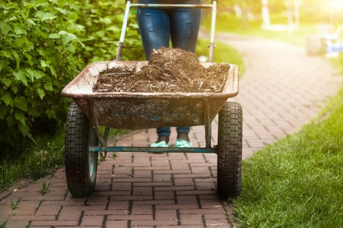 A person standing behind a metal wheelbarrow filled with dark brown soil or compost, positioned on a brick pathway within a residential garden or driveway. The wheelbarrow has a single wheel at the front and two supporting legs at the rear, with signs of dirt and weathering on its surface. The individual is wearing blue jeans and dark shoes, with their upper body not visible in the image. To the left, a green hedge lines the edge of the pathway, while on the right, a strip of trimmed grass borders the bricks. In the background, part of a paved area and a sunny, well-maintained garden scene are visible, suggesting an outdoor setting suitable for rubbish removal or garden clearance activities, with the context aligning with services offered by Commercial Waste Archway in North London.