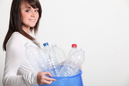 A young woman with shoulder-length brown hair and light skin is smiling and looking towards the camera while holding a blue plastic container filled with multiple discarded empty plastic bottles, including clear and transparent bottles with varied red and blue caps. The bottles are all made from PET plastic and are loosely arranged, some leaning against others inside the container. She is wearing a white long-sleeved top and stands against a plain white background, which provides a clean, professional setting. The image focuses on waste collection, illustrating proper disposal of plastic bottles, relevant to rubbish removal services such as those offered by Commercial Waste Archway in the North London area, including Archway postcodes. The bright lighting and simple environment highlight the clean, straightforward approach to waste management practices, emphasizing environmentally conscious disposal and recycling efforts in urban settings.