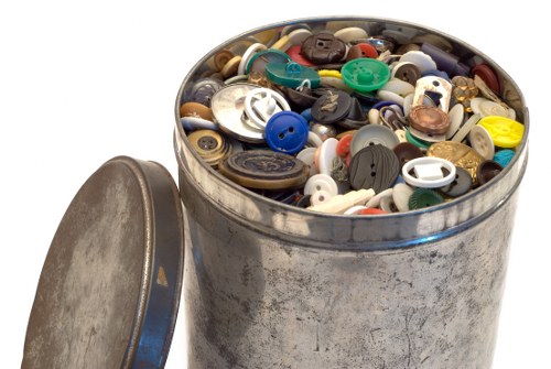 A round metal container with a partially removed lid, filled to the brim with a variety of used plastic and metal buttons of different sizes, shapes, and colours. The buttons include blue, black, white, beige, yellow, green, and brown, with some metallic or shiny finishes. The container appears to be situated outdoors on a paved or concrete surface, possibly in a garden or near a building, with natural daylight illuminating the scene. The overall environment suggests a setting where waste, including miscellaneous clothing fasteners or craft supplies, is being collected or disposed of, consistent with rubbish removal services provided by Commercial Waste Archway in the north London area.