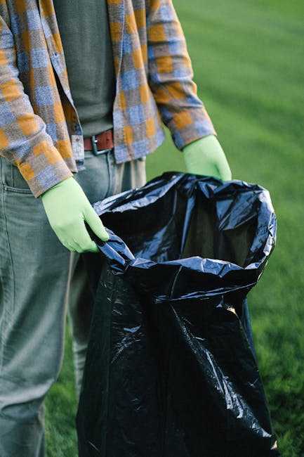 A person wearing a plaid shirt in shades of yellow, gray, and blue, along with gloves, is standing outdoors on a grassy area and holding open a large black rubbish bag made of thick plastic material. 