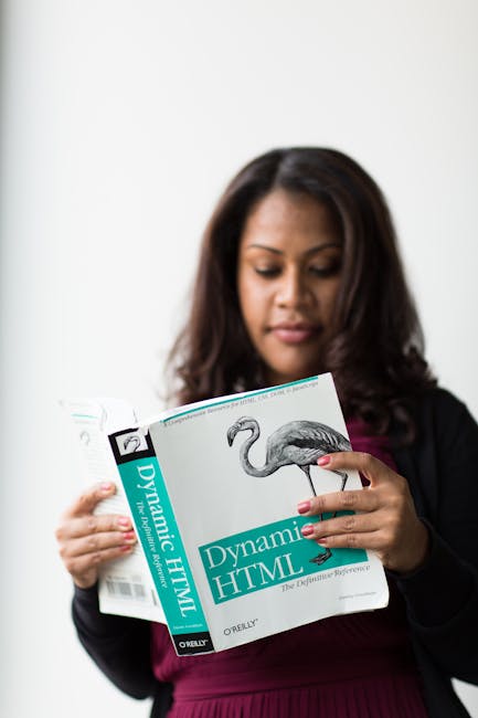 A person wearing a plaid shirt in shades of yellow, gray, and blue, along with gloves, is standing outdoors on a grassy area and holding open a large black rubbish bag made of thick plastic material. 