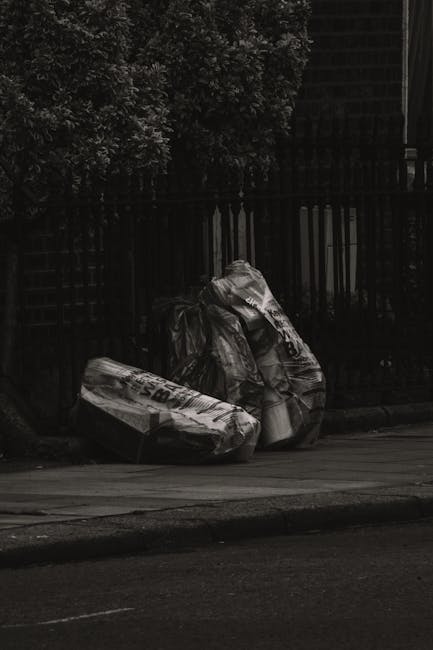 Two large black trash bags made of thick, crinkled plastic are stacked against a black metal fence on a pavement. The bags appear to contain household or garden waste, with some parts of the plastic s