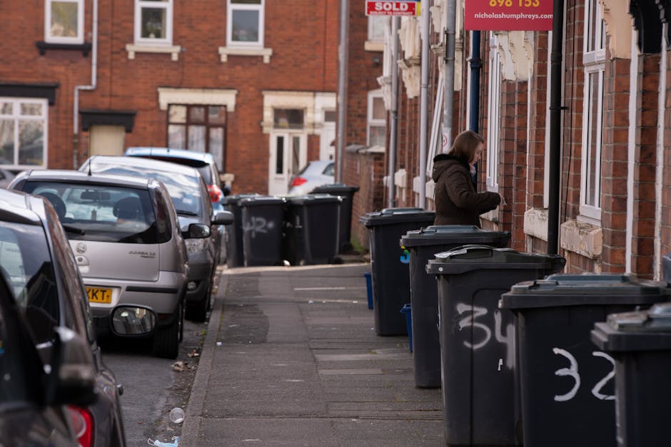 Two large black trash bags made of thick, crinkled plastic are stacked against a black metal fence on a pavement. The bags appear to contain household or garden waste, with some parts of the plastic s